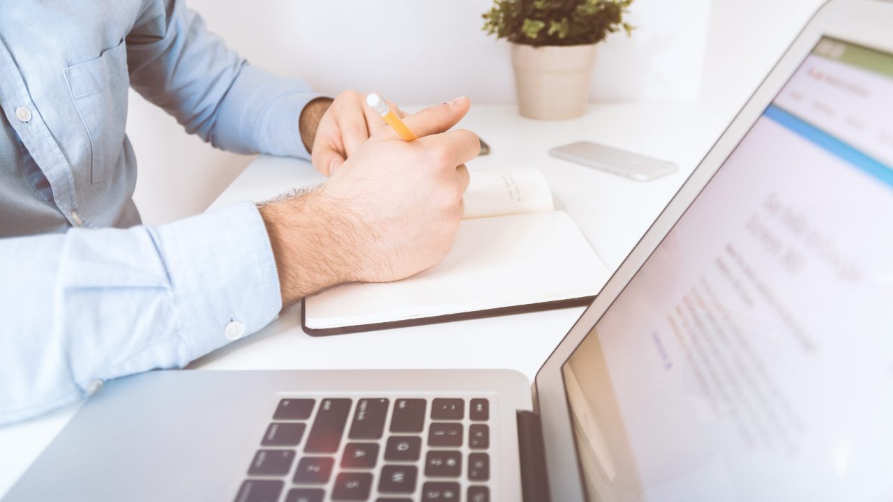 Man takes notes while accessing an online resource on a laptop