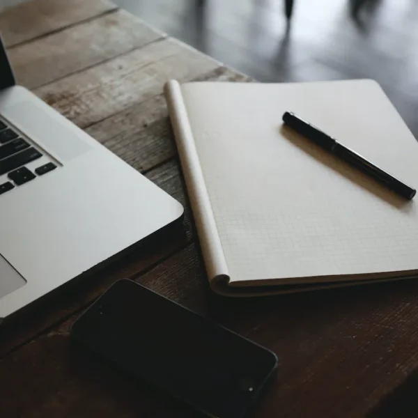 Laptop, notebook with pen, and smartphone on a wooden table, creating a productive workspace vibe.