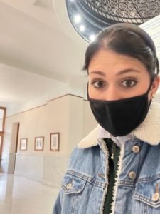 Grace Clucas stands in the hallway of a brightly lit building. She is wearing a mask and her hair is tied back and she is wearing a denim jacket. 