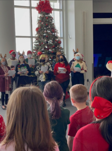 A group of half a dozen adults wearing Santa hats and reindeer antlers sings for a group of small children. A Christmas tree sparkles merrily in the background. 